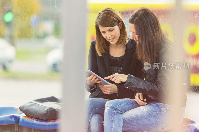 Two friends using digital tablet on bus station